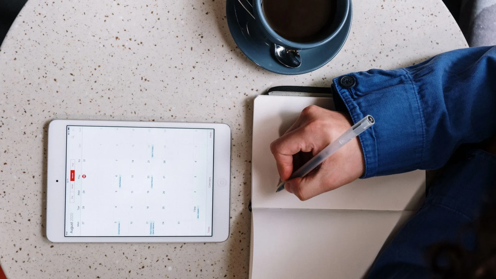 Hand writing in a notebook beside a tablet displaying a calendar